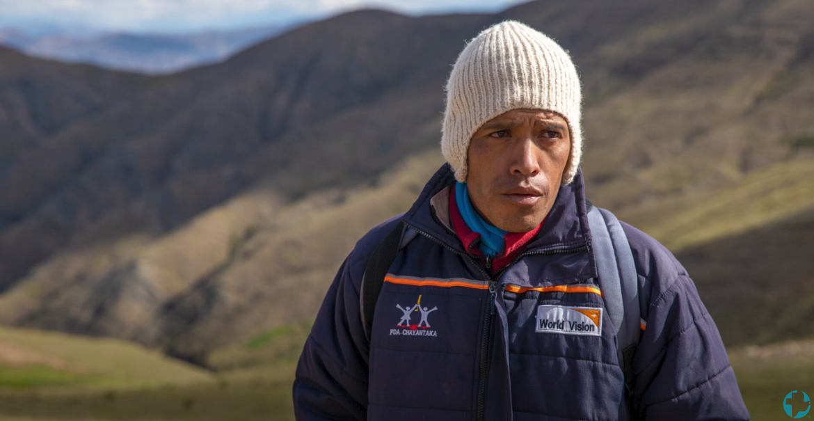 A Bolivian man wearing a blue jacket and a white hat that covers his ears. Mountains are in the background.