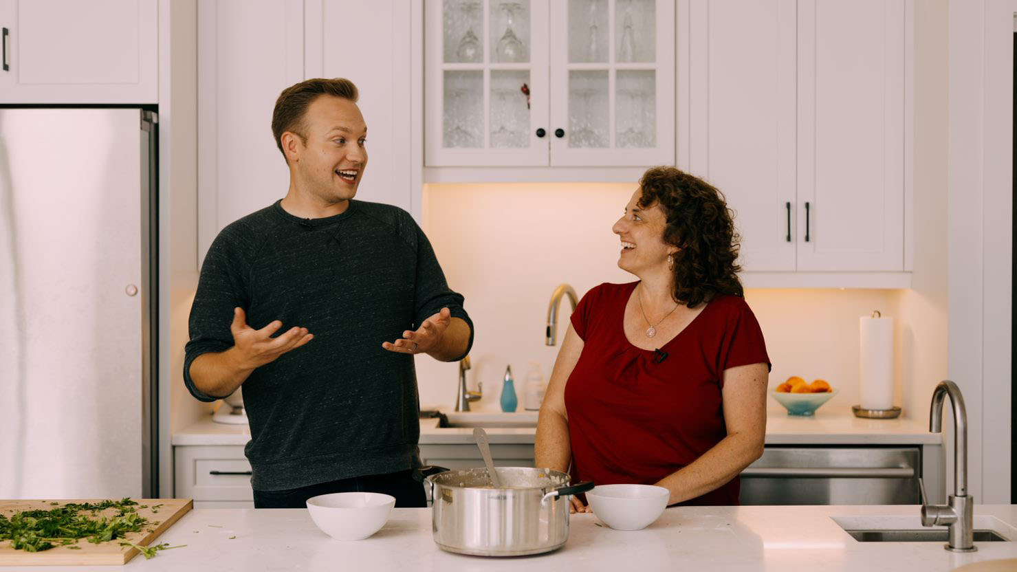 A man and a woman stand behind a kitchen counter and talk