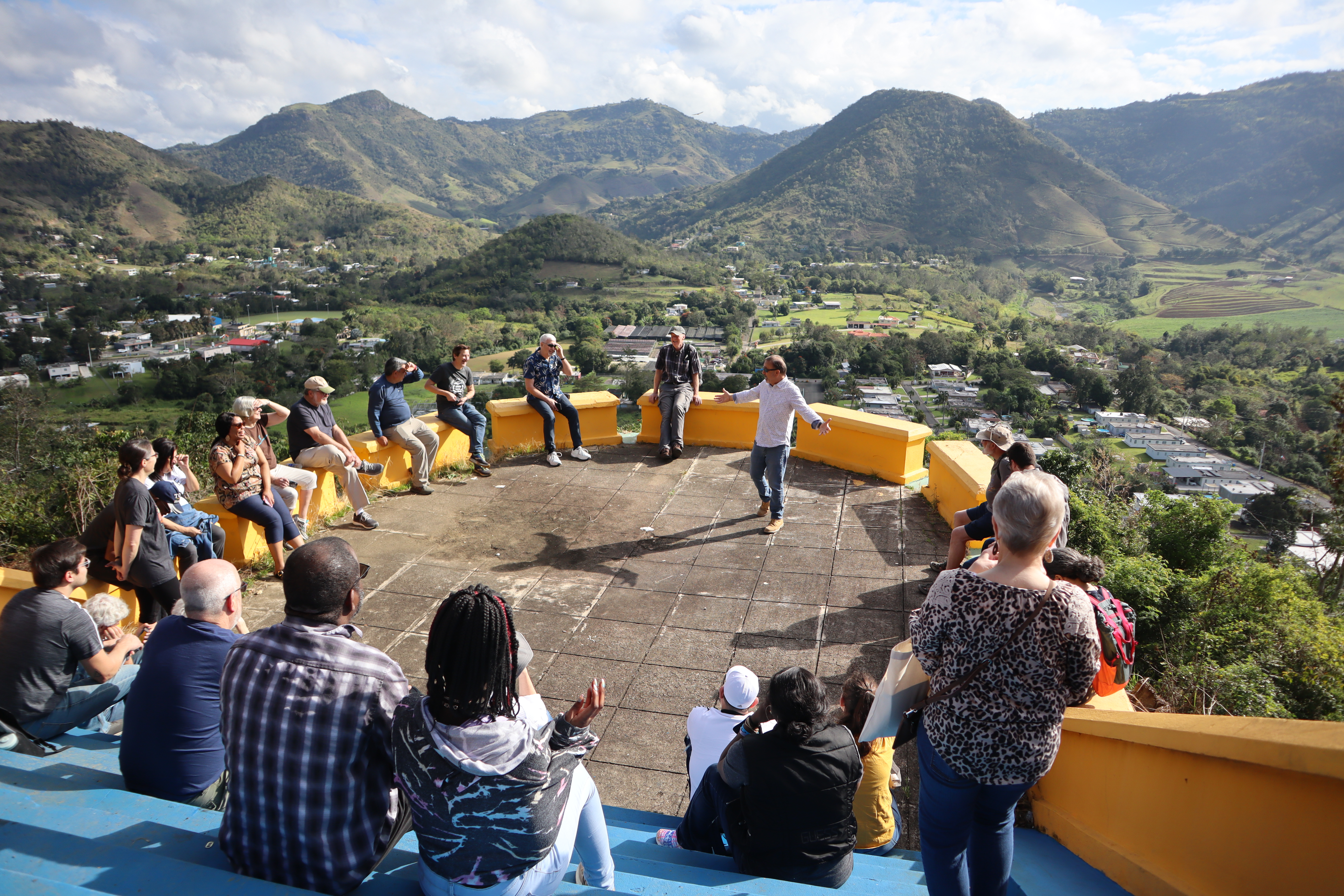 A group of people sitting in a circle listening to a speaker surrounded by grassy hills