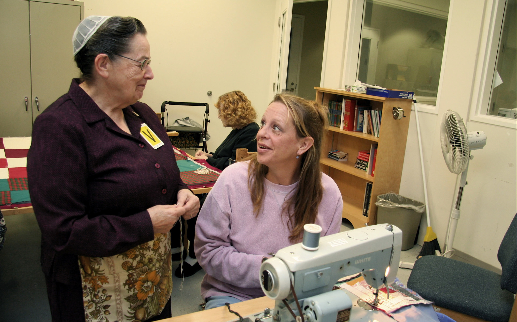 A woman stands next to a woman sitting at a sewing machine