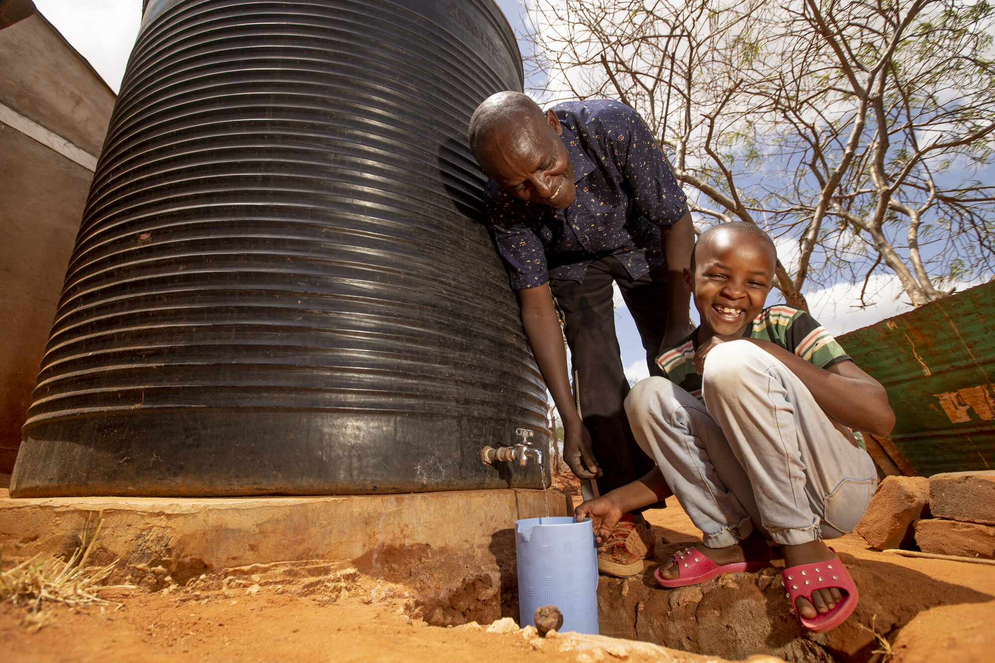 A father and son collect water from a large outdoor rain barrel