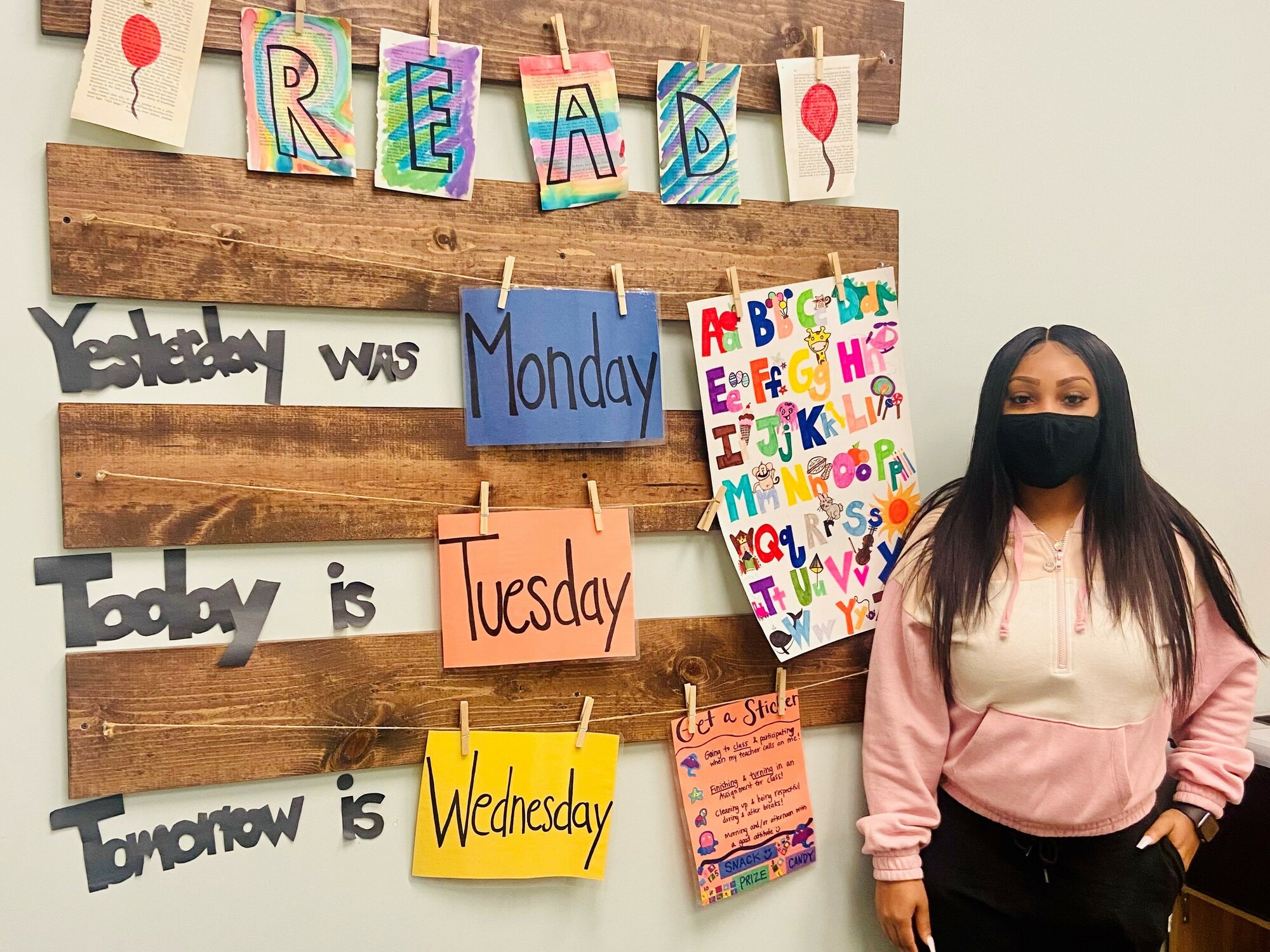 A young woman in a face mask stands next to a wall covered with colorful learning materials in a classroom