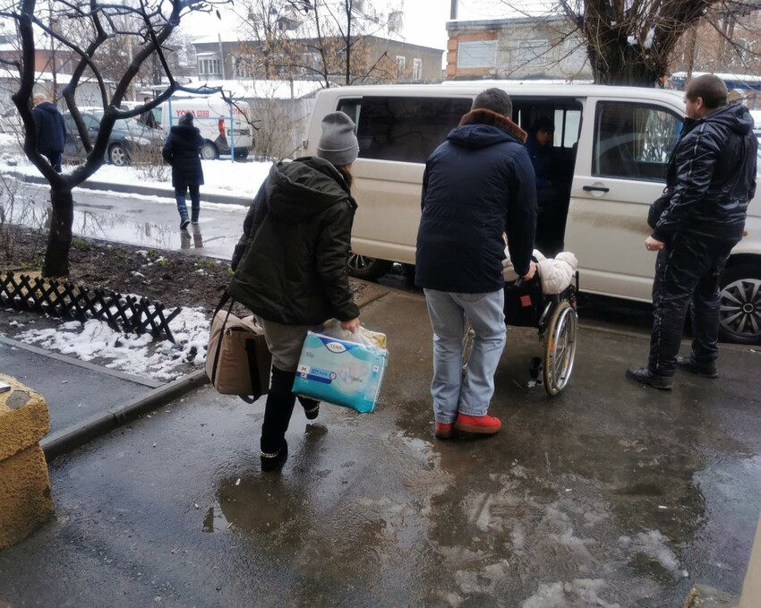 Three people on a sidewalk are heading towards a white van. One is carrying bags, while one is pushing the other in a wheel chair. 