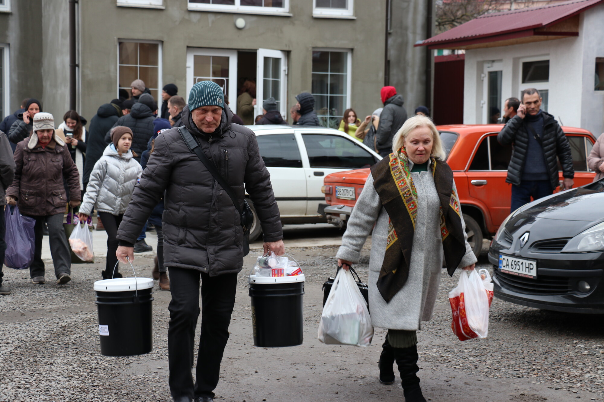 A man and a woman walk away from a building caring bags and five gallon buckets