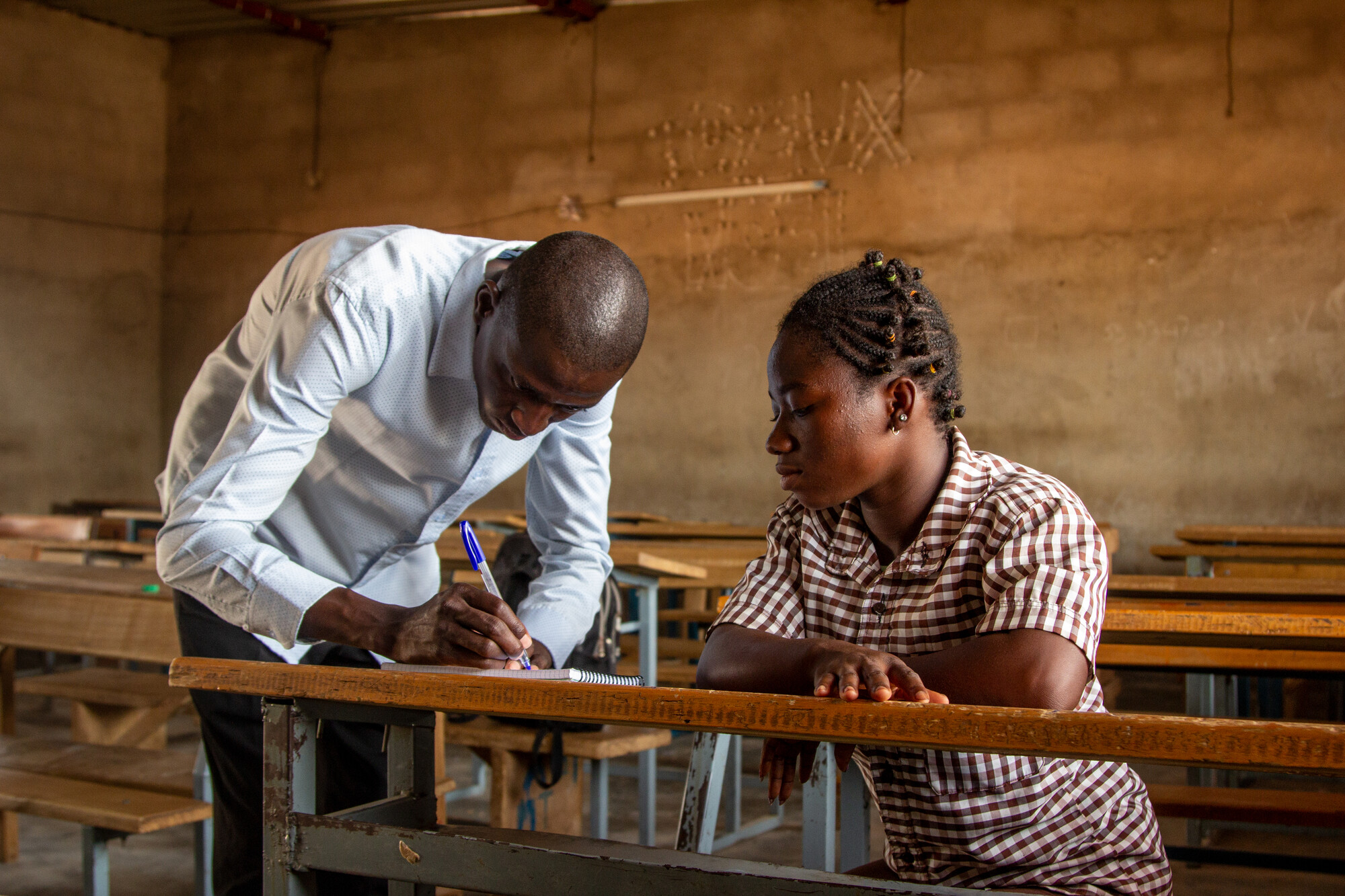A teacher leans over a desk and writes something as a student looks on.