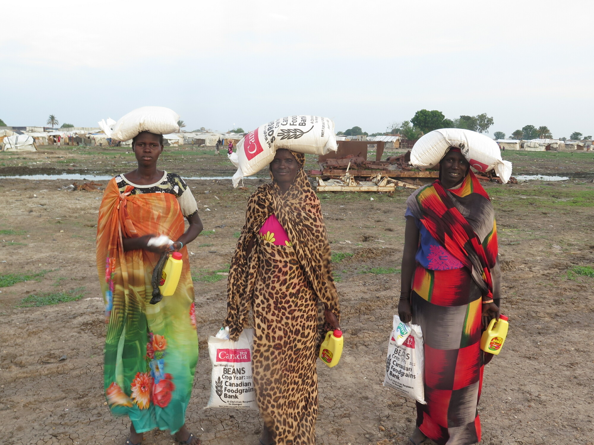 Three South Sudanese woman hold food supplies in their arms and on their heads.