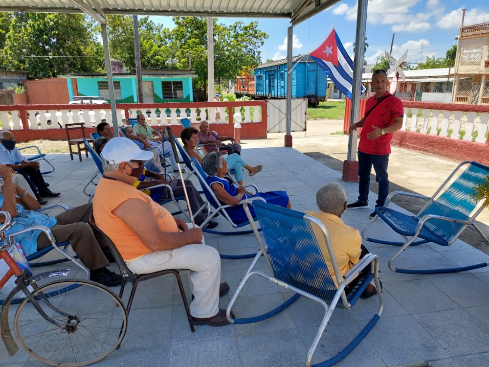 A man in a red shirt speaks to a group of elderly Cubans who are sitting in front of him.