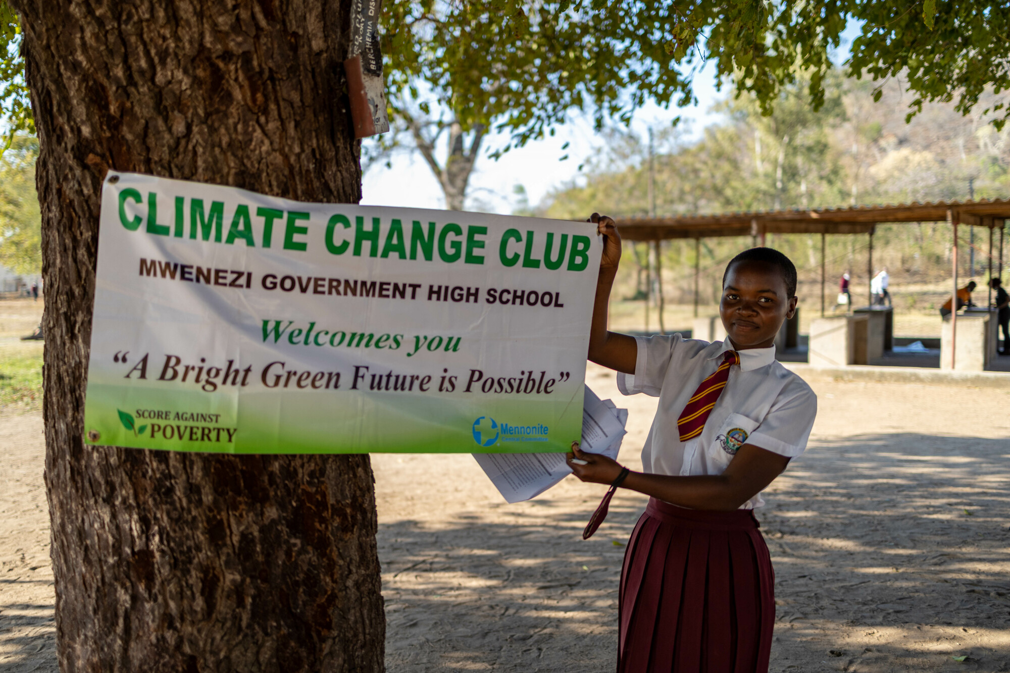 A student in a school uniform holds up one end of a banner. (The other half is attached to a tree trunk). The banner says, "Climate change club. Mwenezi Government High School Welcomes you. 'A Bright Green Future is Possible.'"