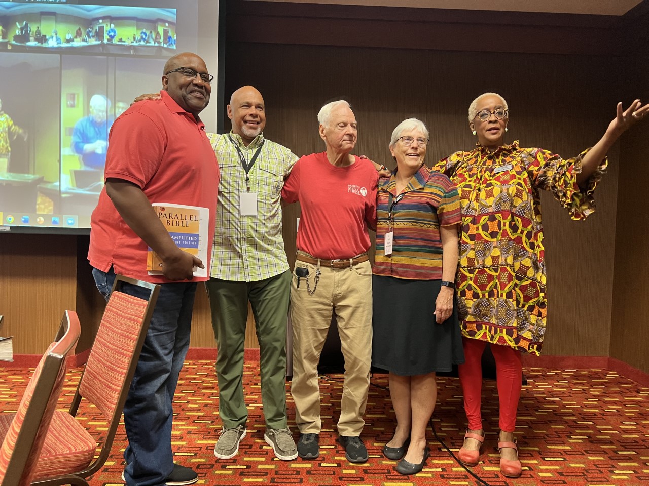 Spencer Lindsay, left, and Jerry Horn (both wearing red shirts) from Working Men of Christ stand with U.S. board member Terry Hunt (green shirt) and MCC U.S. Executive Director Ann Graber Hershberger (black skirt) and MCC U.S. Central States Executive Director Michelle Armster, right, after their presentation at the October 2022 MCC U.S. board meeting.
