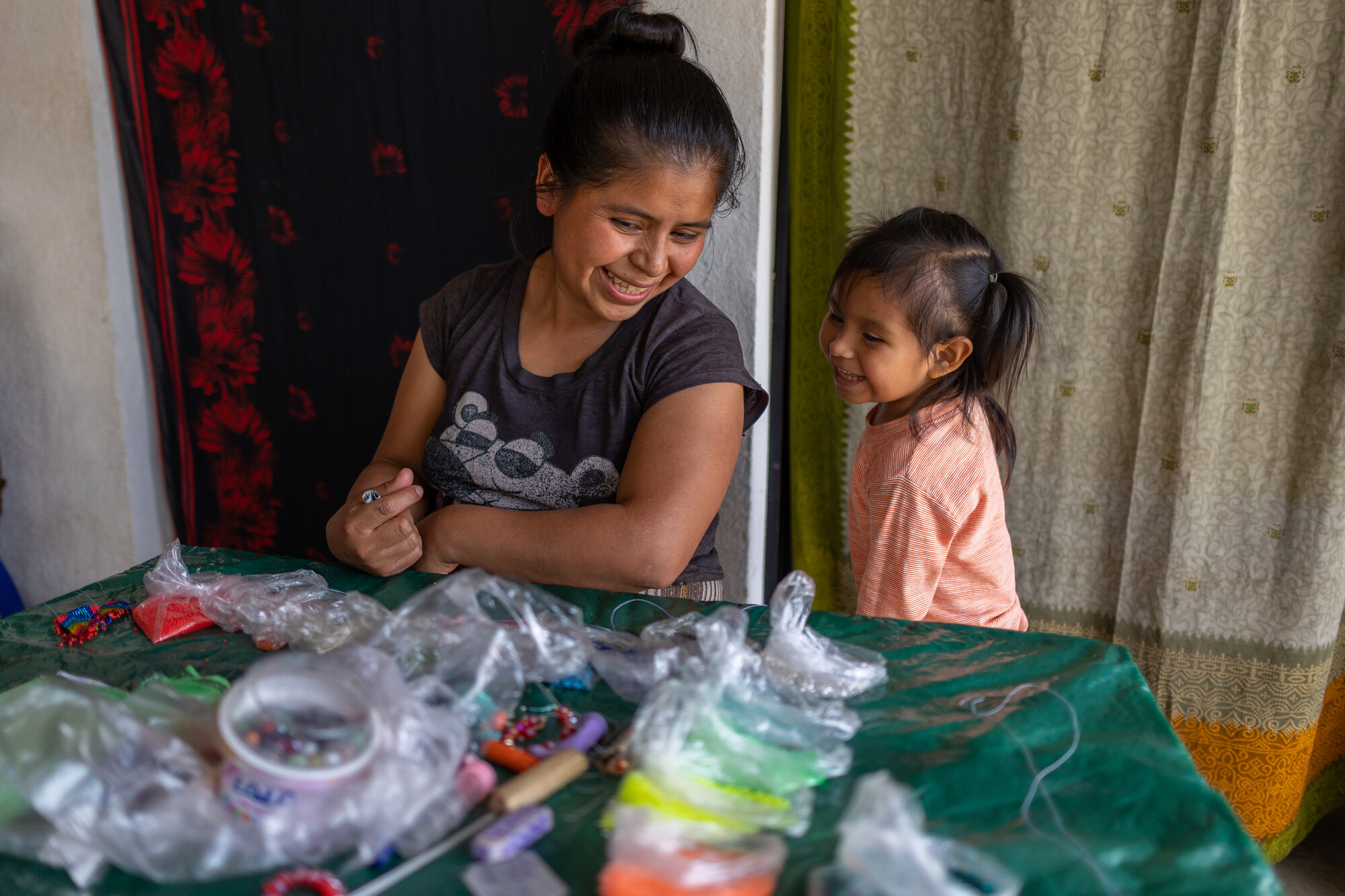 A woman and child smile at each other. The woman is sitting at a table full of  beadwork