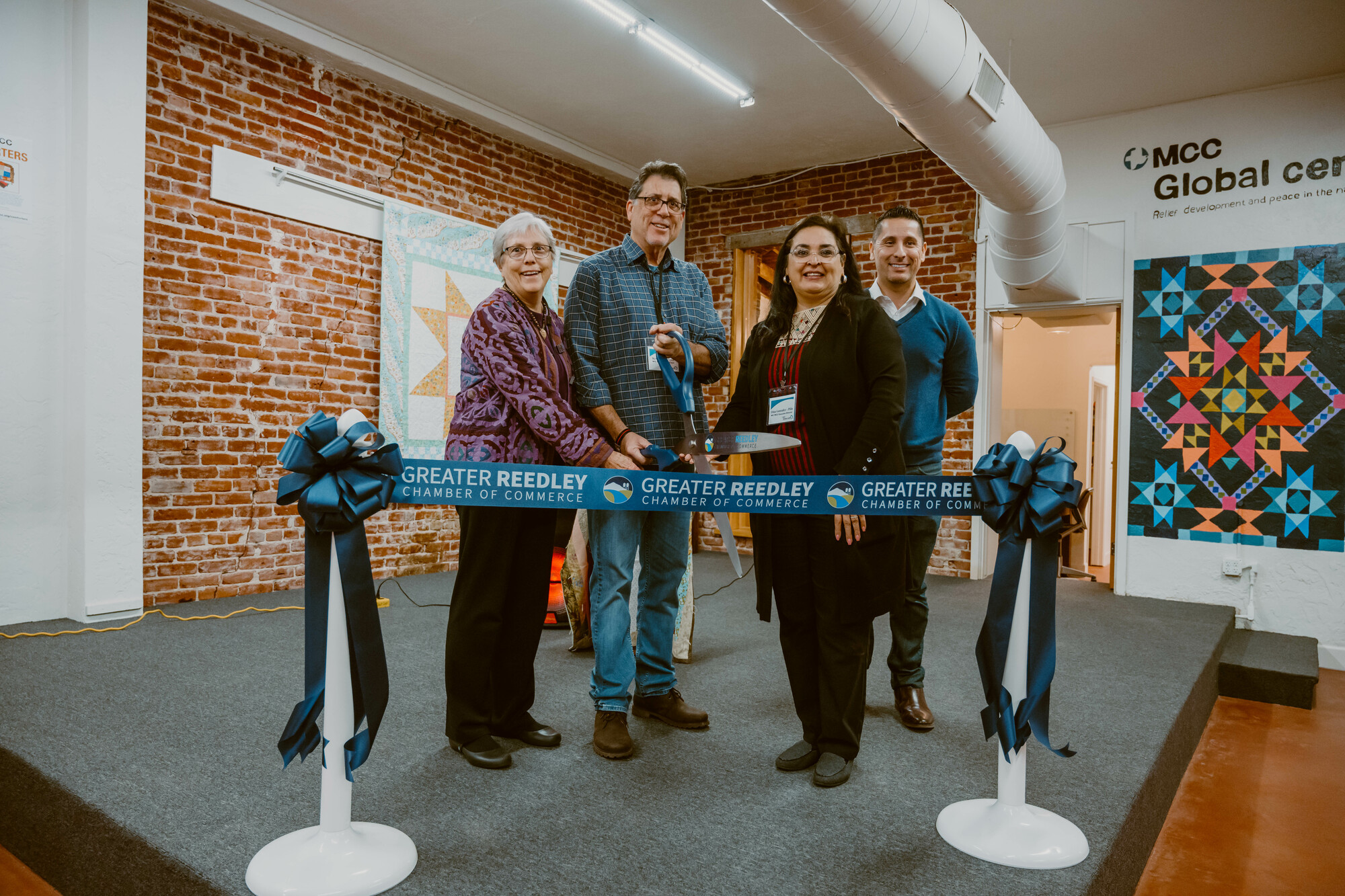 Four people stand behind a blue ribbon. Two people hold on to a large pair of scissors 