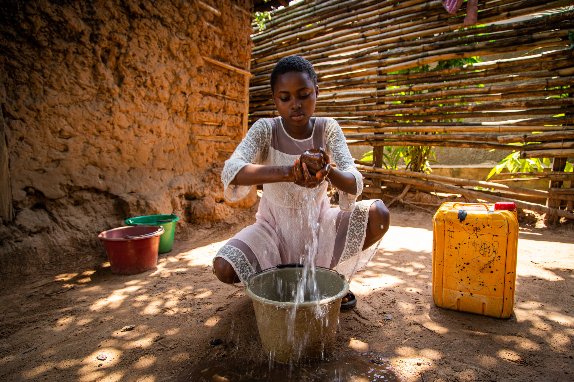 A girl in a white dress washes her hands in a bucket. She has a stream of water falling off her hands.