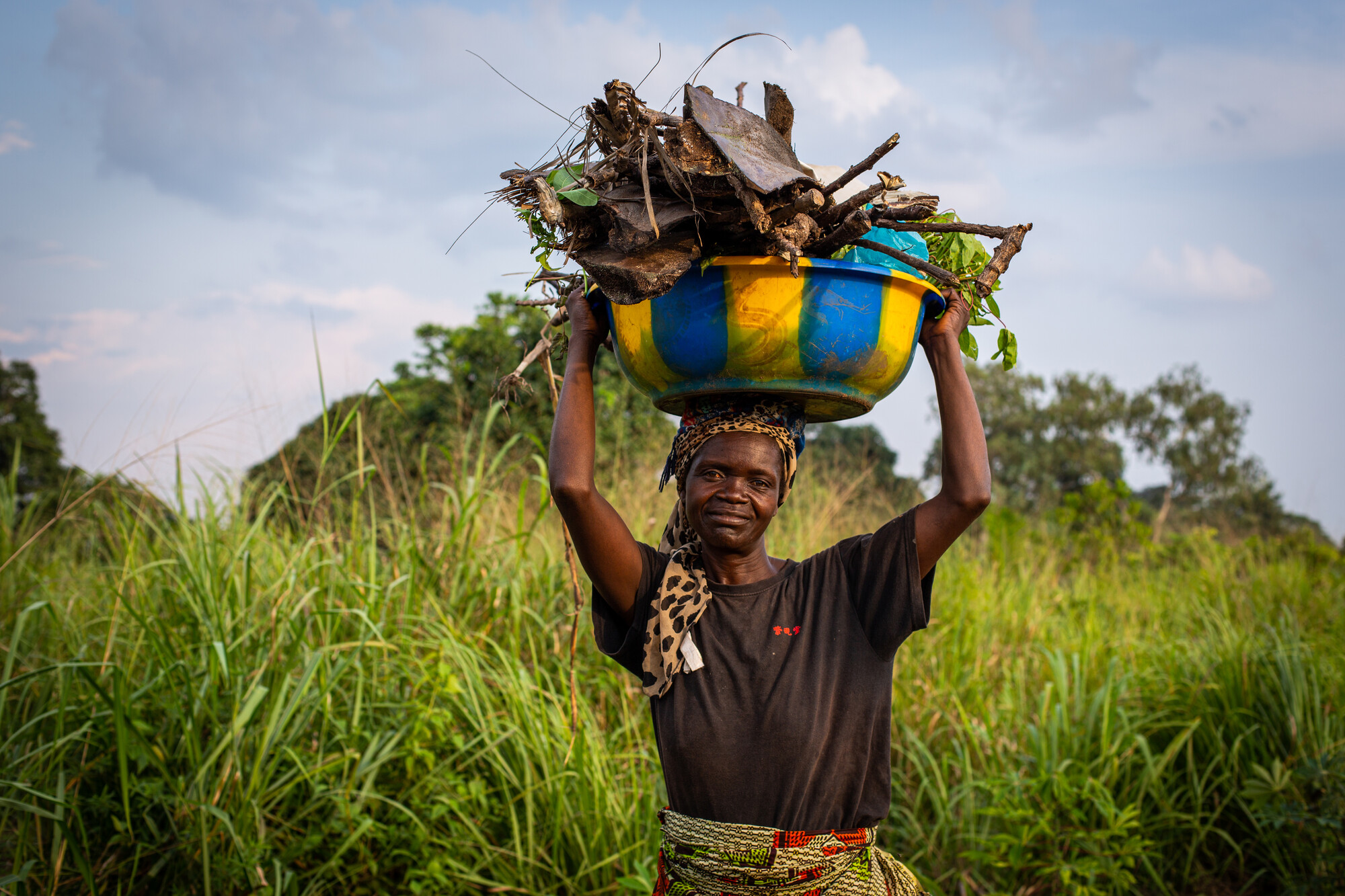 A woman holds a large yellow and blue bucket on her head. The bucket is full of firewood.