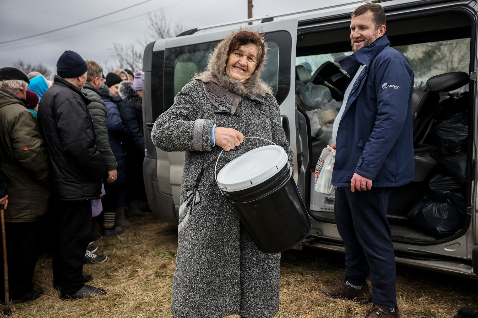 A woman in a winter coat holds up a five gallon bucket and smiles at the camera.