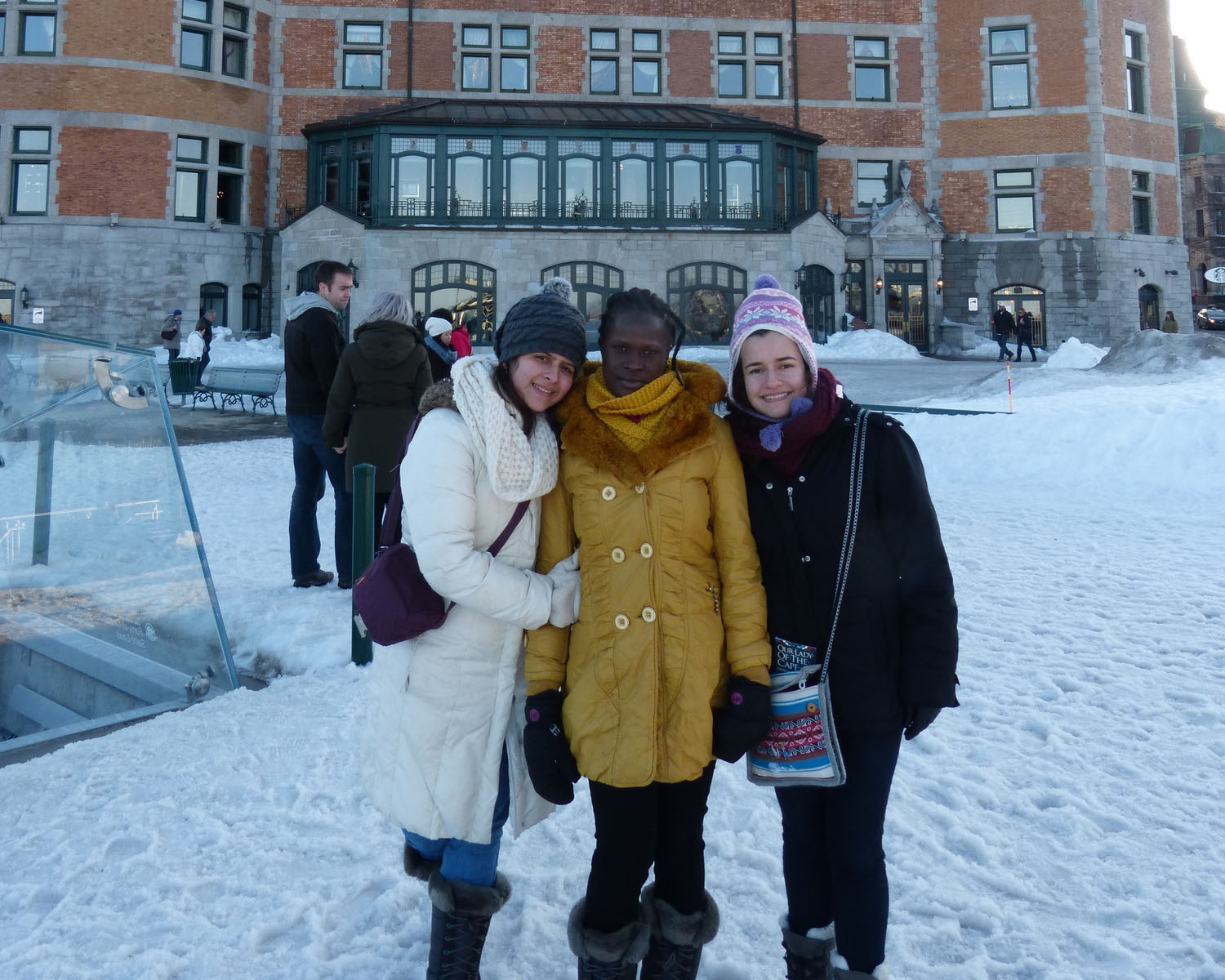 Three people in winter coats stand in the snow in front of a city building