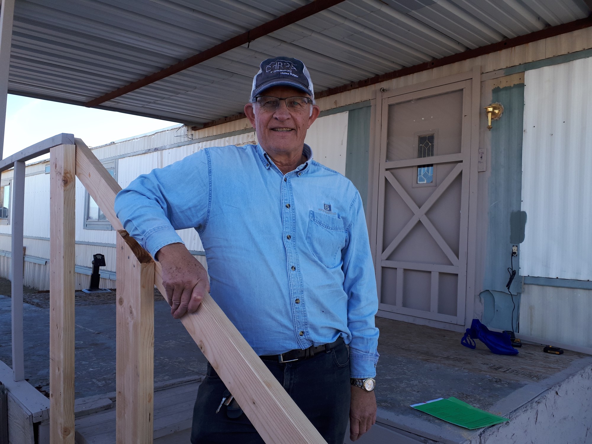An older man in a ball cap stands beside a wooden railing on a mobile home