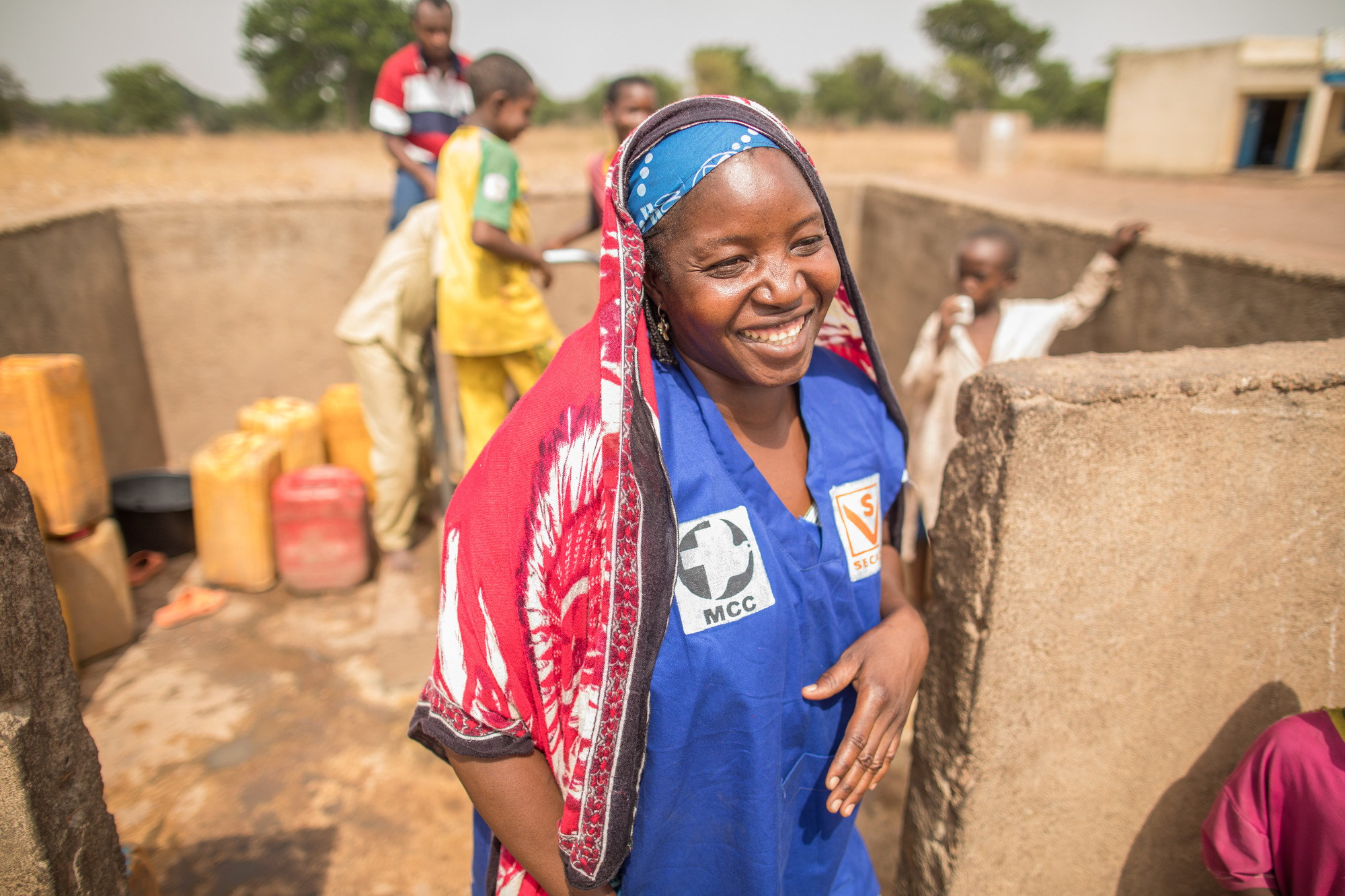 A Chadian woman in a blue shirt and red scarf covering her read laughs near a well.