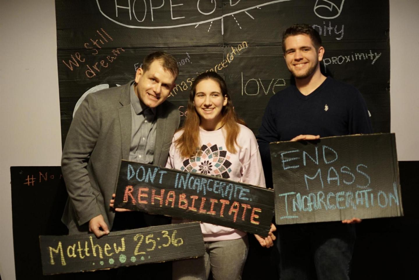 A group of three people pose in a makeshift photo booth with signs that say, "Matthew 25:26," "Don't incarcerate. Rehabilitate," and "End mass incarceration"