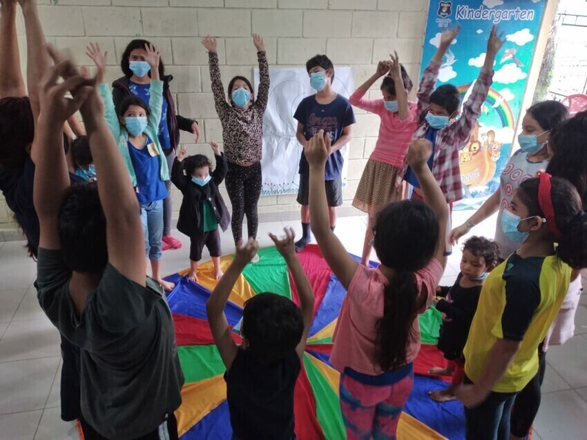 A group of children stand around a colorful parachute on the ground. They are raising their hands in the air.