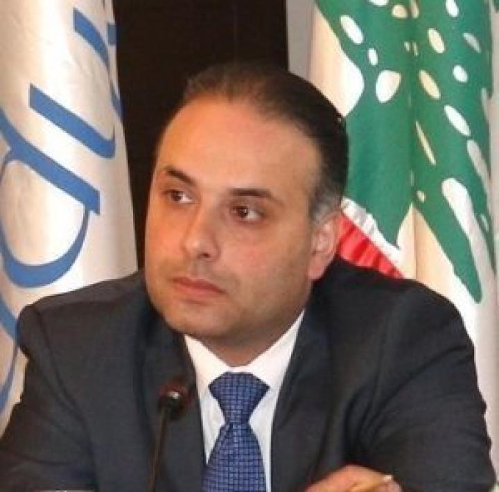 A man wearing a black suit and blue tie sits in front of two flags. 