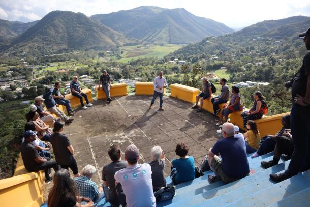 An overview shot of a large group of people sitting in a circle listening to someone speak. Behind them are the mountains of Puerto Rico