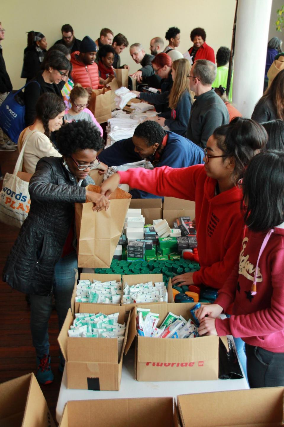 A large group of people standing around a long table, working on an assembly line putting hygiene items into brown paper bags.