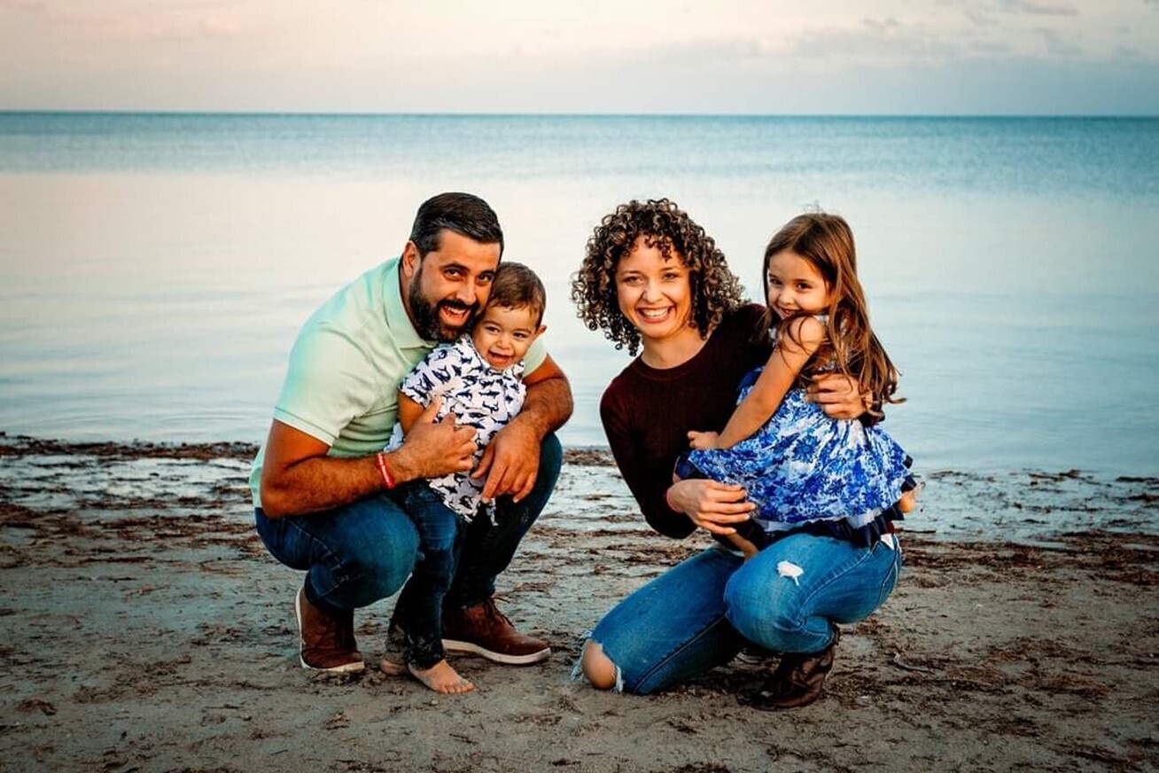 A family of four kneal together in the sand at the beach. The ocean is behind them.