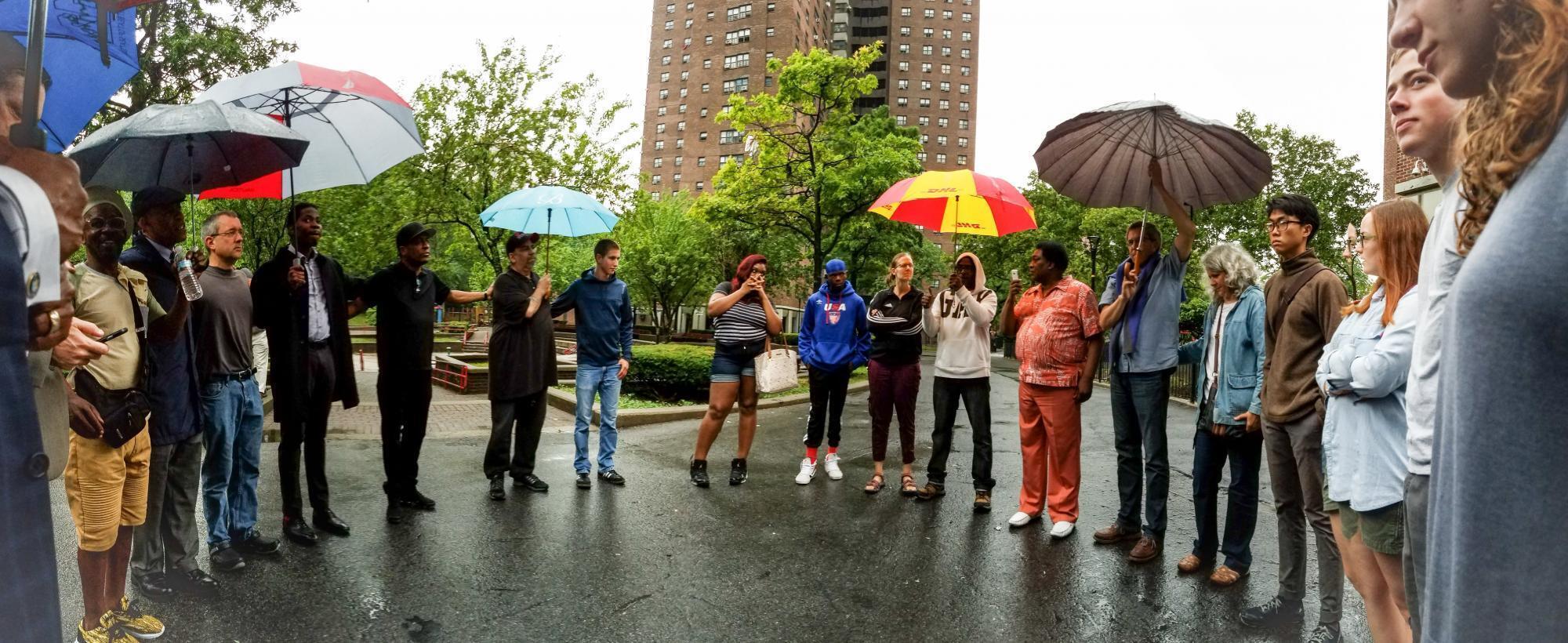 A group of young adults stand on pavement in a circle. Many of them are holding umbrellas.