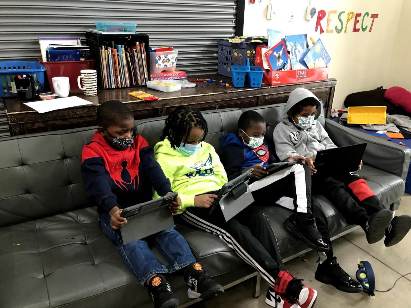 Four kids sit on a couch wearing face masks and looking at tablets