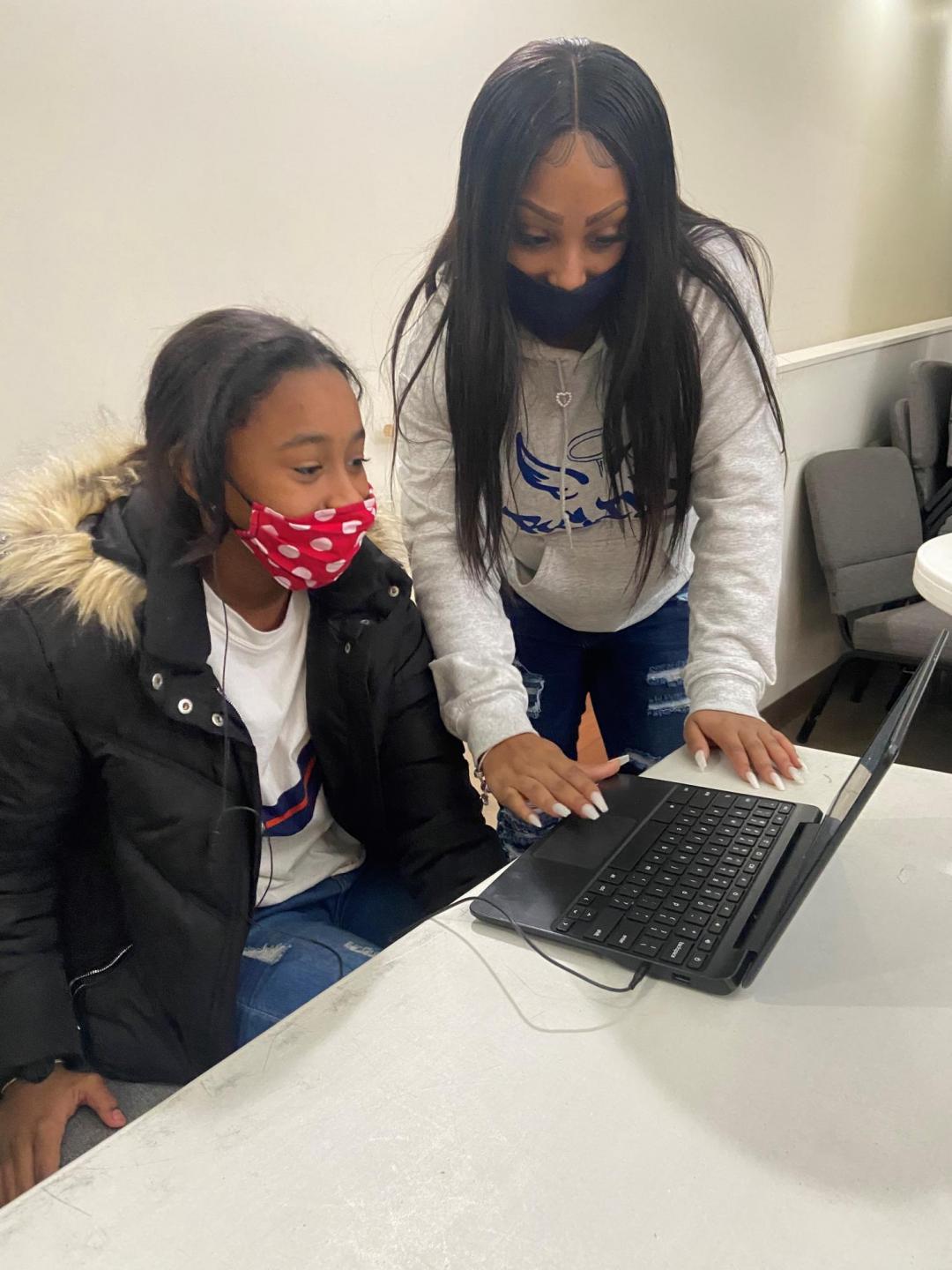 A young adult leans over a lap top and assist a child who is sitting in front of the computer