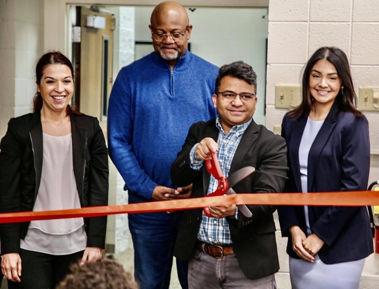 Four people stand behind a red ribbon. One man has a giant pair of scissors and is getting ready to cut the ribbon.