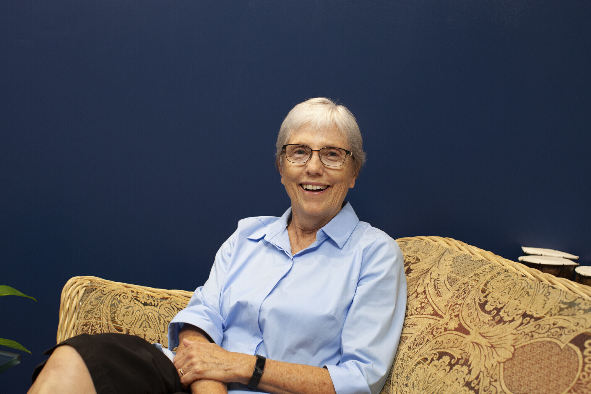 A smiling woman with glasses and short silver hair, wearing a blue collared button up shirt, sits on a couch and looks to her left into the camera.