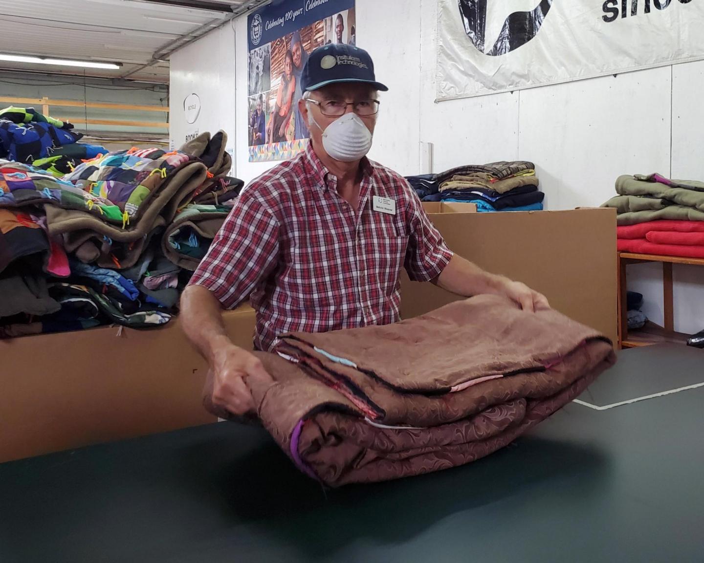 A man in a ball cap and a face mask holds a folded comforter in a warehouse.