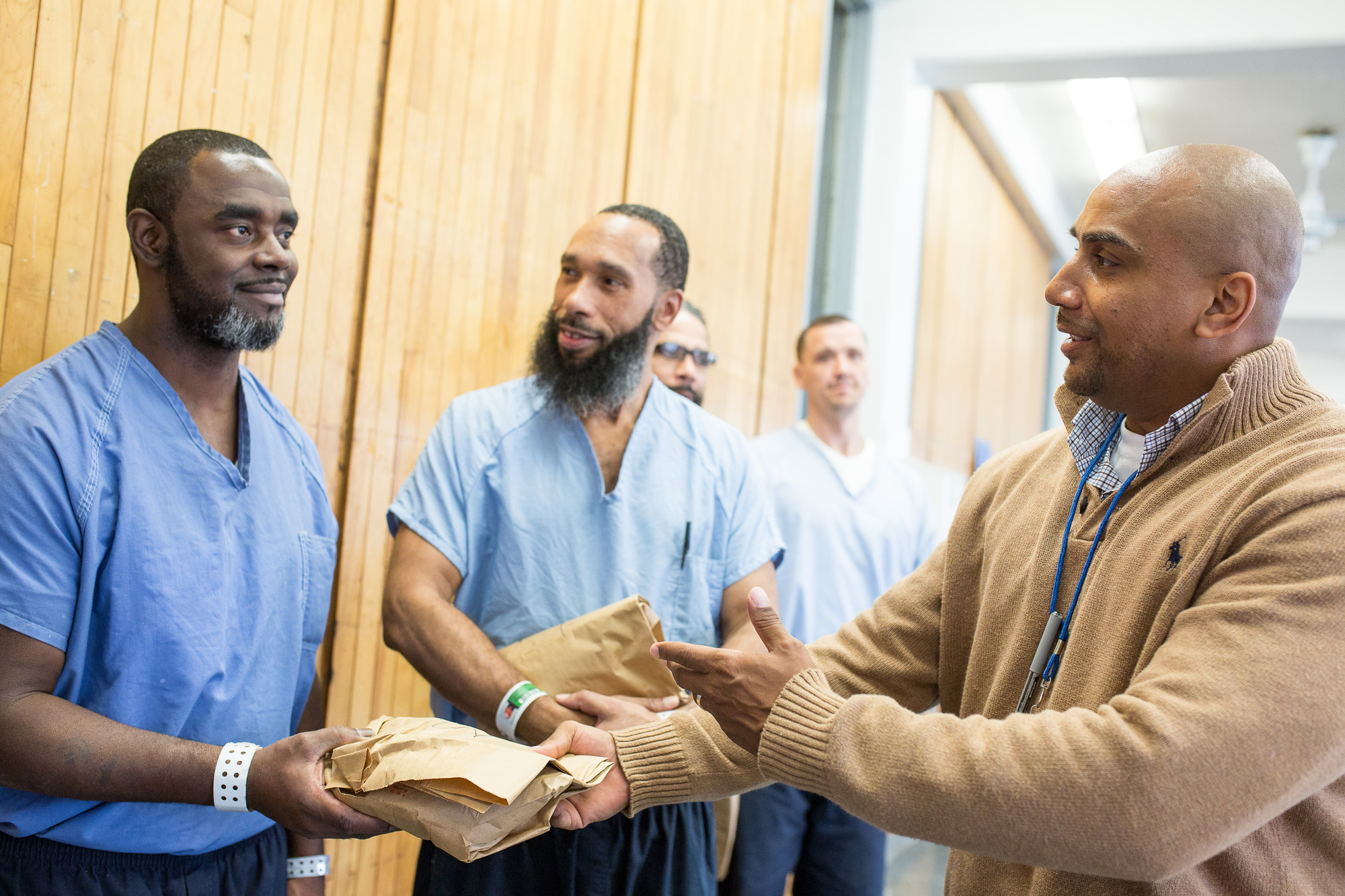 A man in a tan sweater hands a paper bag to another man in blue prison clothes