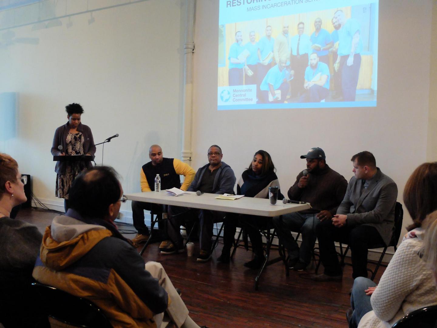 A group of 5 panelist sitting at a table speaking to a audience sitting in front of them. A woman is standing off to the left holding a microphone.