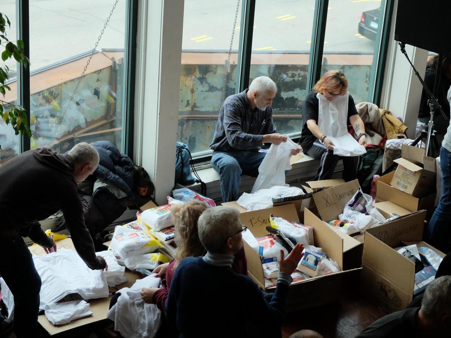 A scattering of people work on folding white t-shirts. There are a bunch of boxes in front of them.