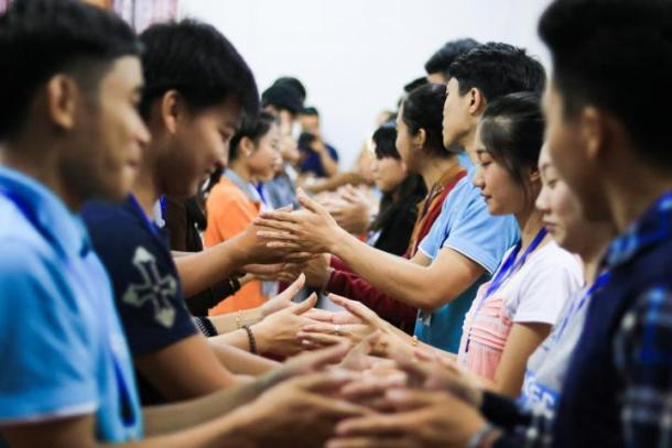 A large group of young adults stand in two rows facing one another with their hands out reached in the middle.