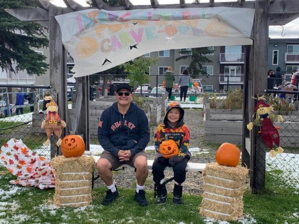 A father and son sit together on a bunch. The son is holding a carved pumpkin