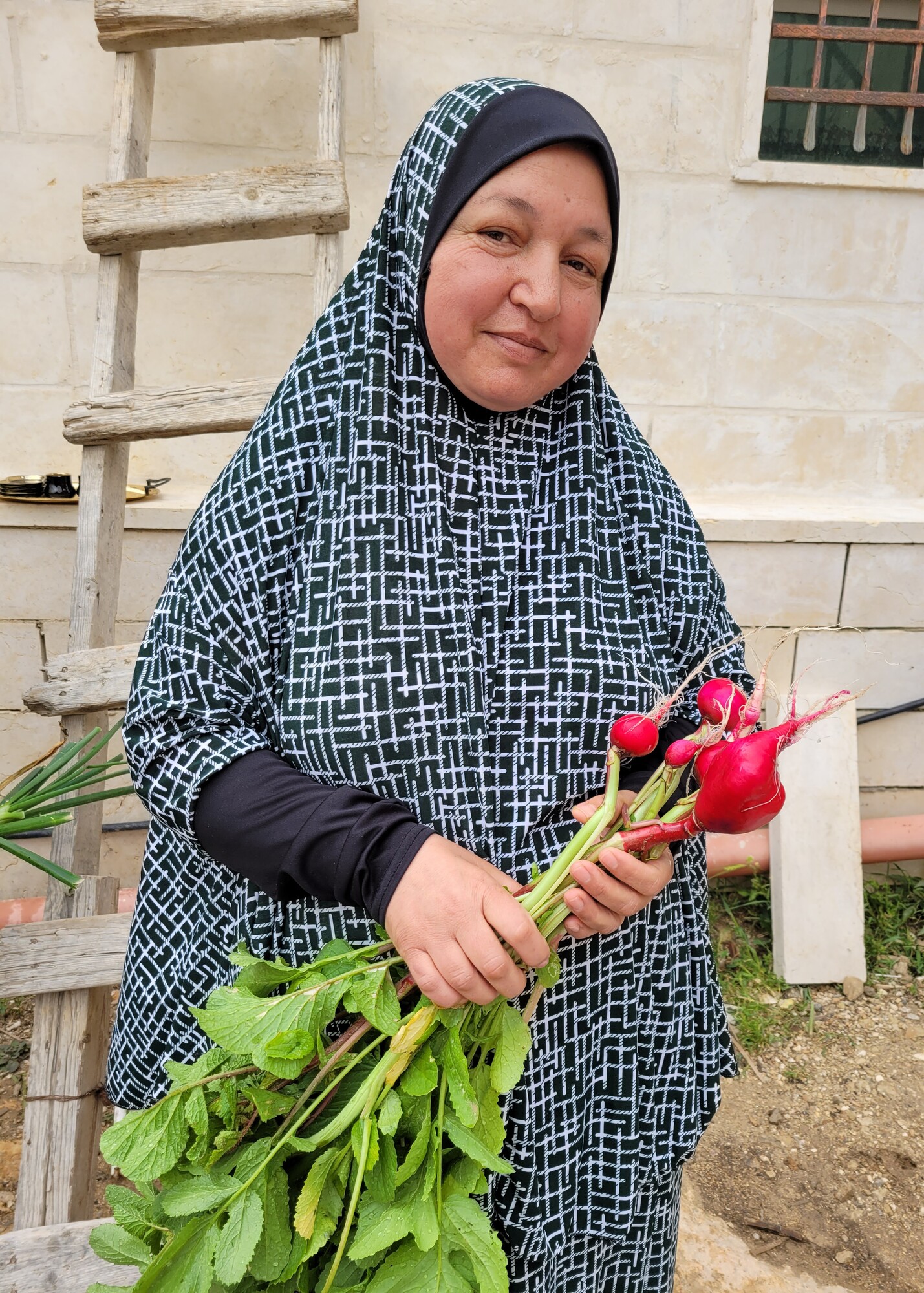 A woman in a black and white hijab stands holding a bunch of radishes