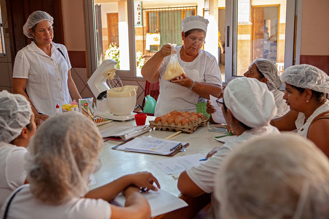 Women wearing white and hair nets gather around a table and watch a cooking instructor