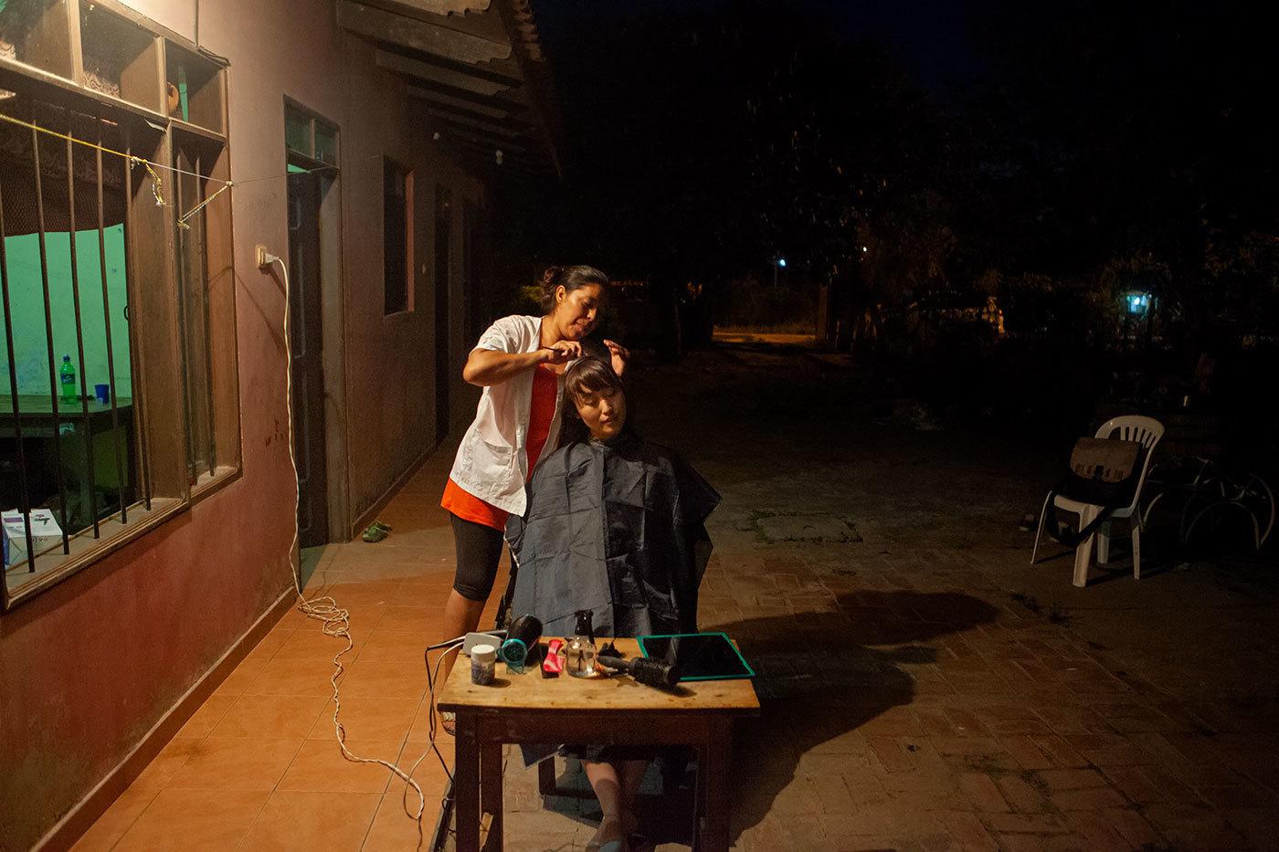 A Bolivian woman cuts another woman's hair outside her home at night