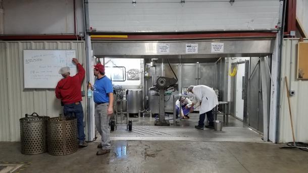 A man writes on a white board while two peopl in white coats and aprons clean a food processing area