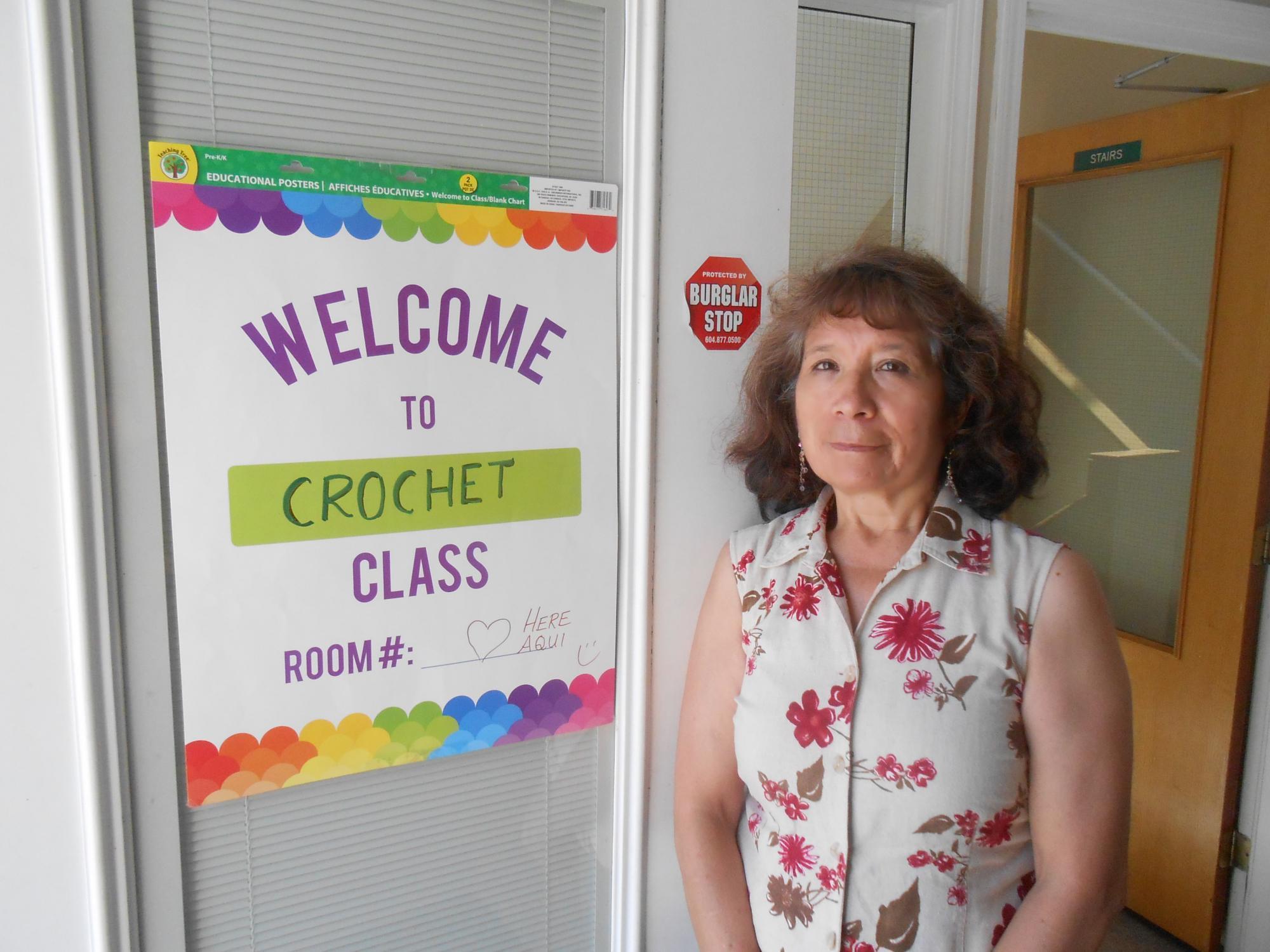 An older woman stands near a sign posted on a window that says, "Welcome to crochet class"