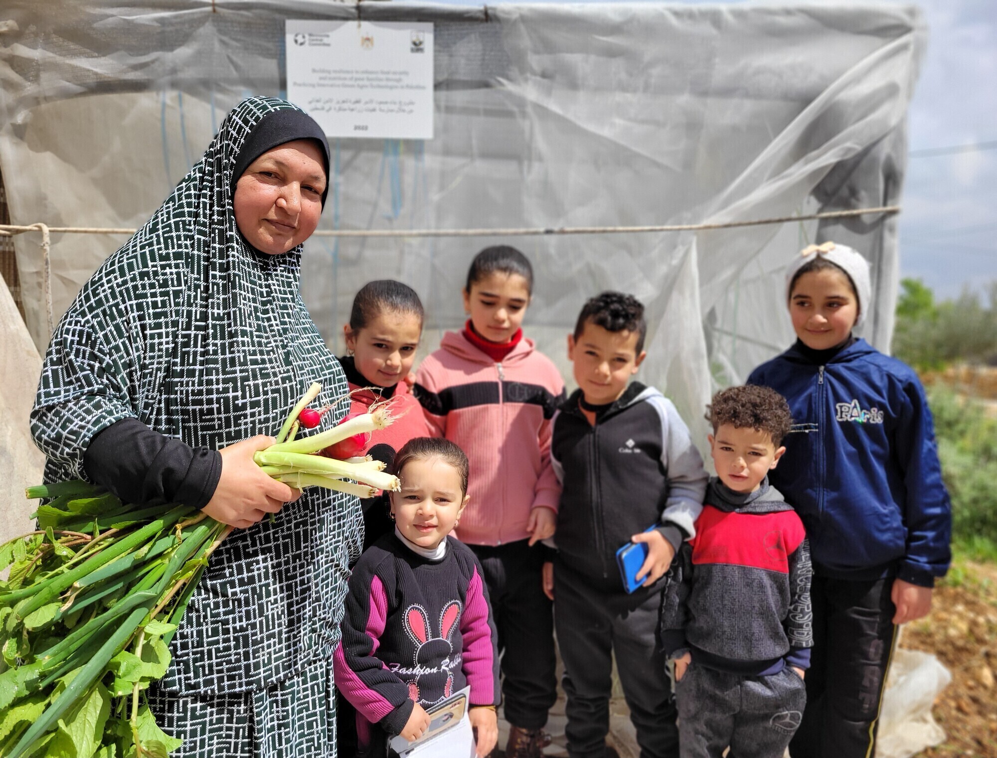 A Palestinian woman stands with six of her grandchildren in a garden. She is holding a bunch of onions