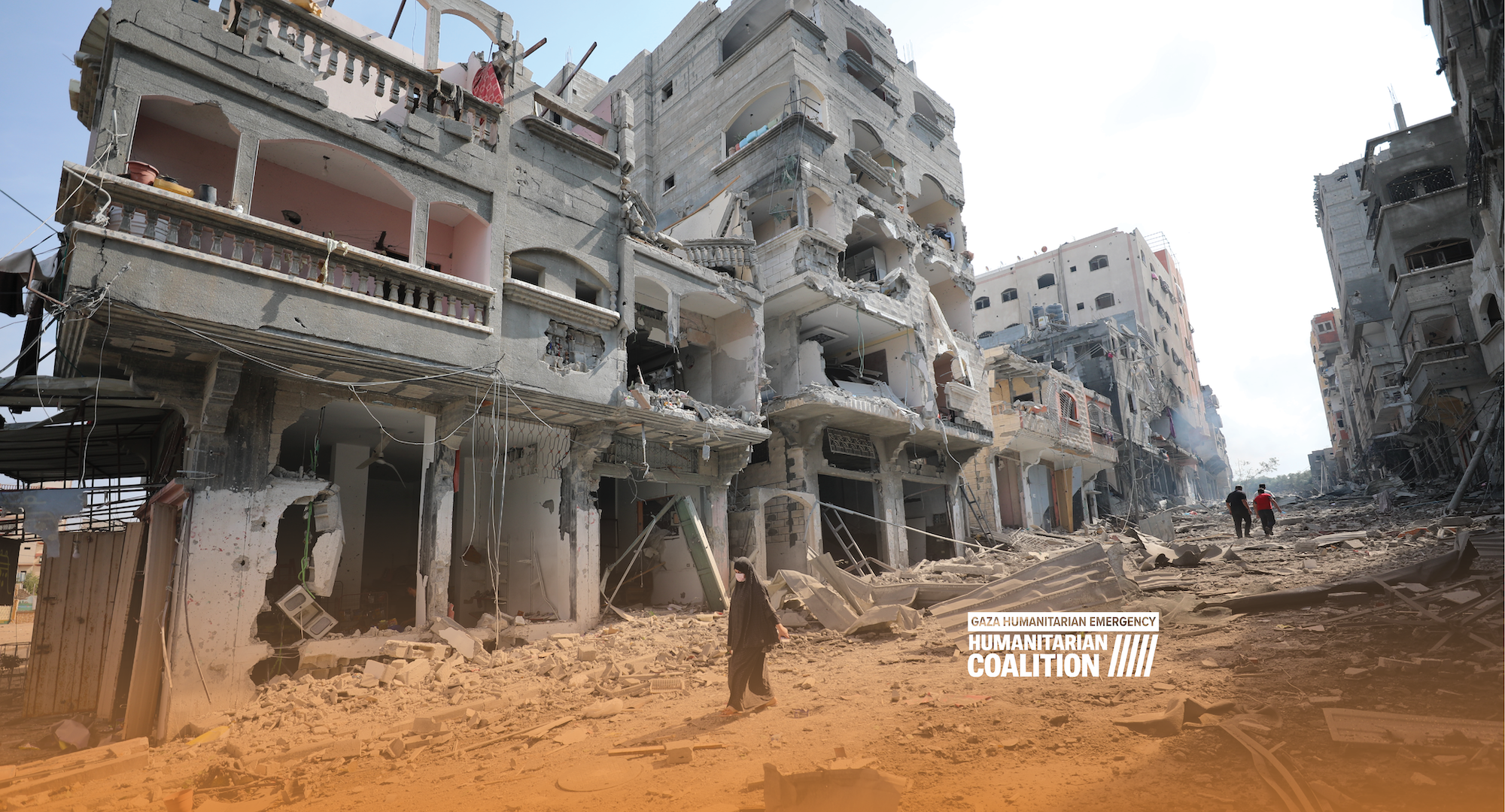 A woman walking by ruined buildings with an overlay of the Humanitarian Coalition logo