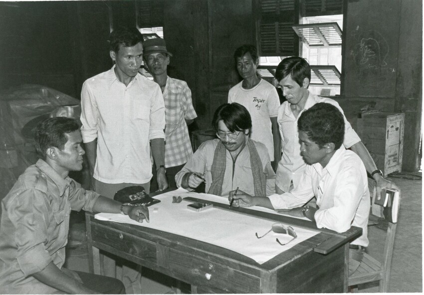 Seven men gather around a desk.