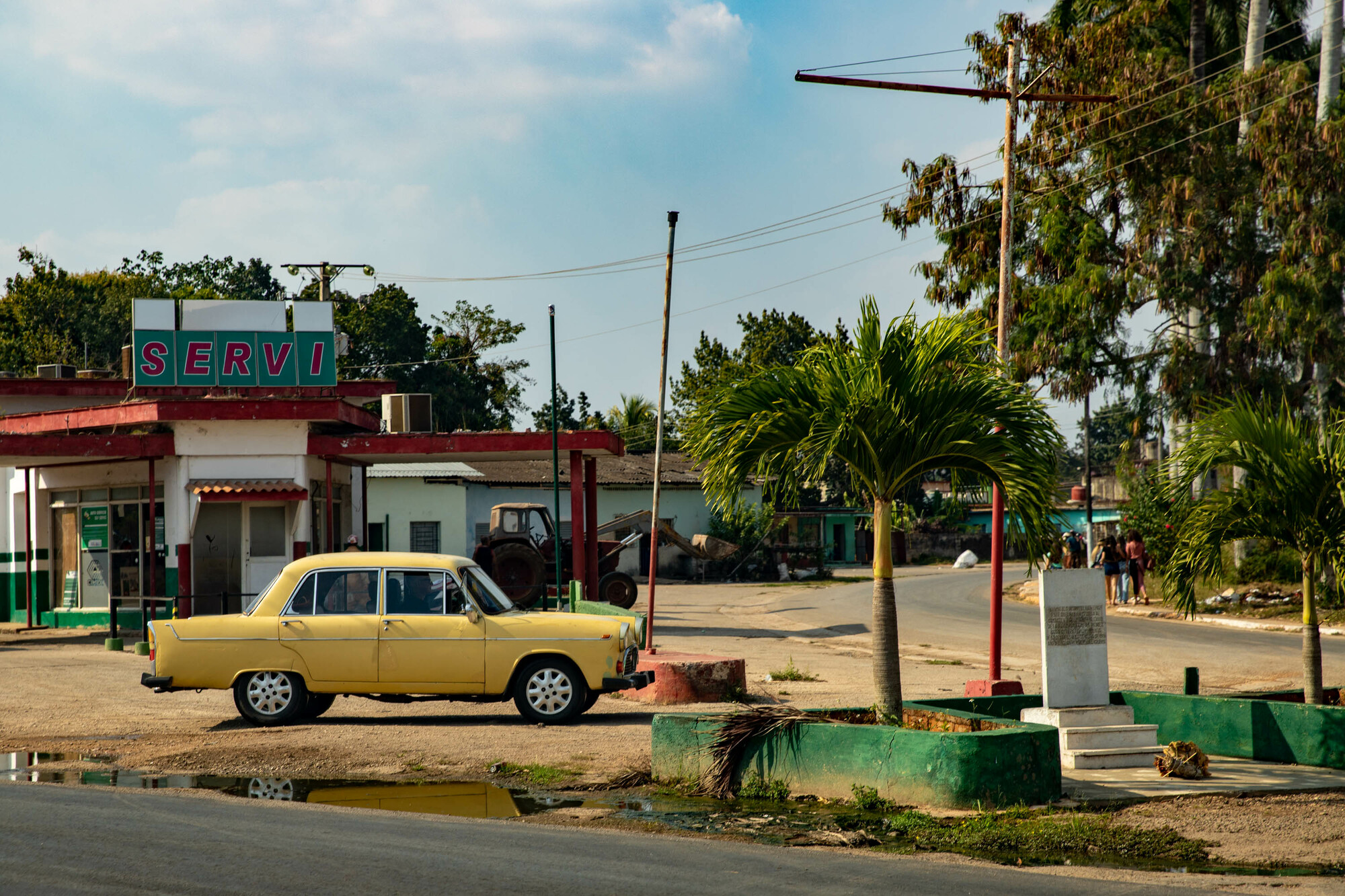 An old yellow car is parked outside of a dated gas station.