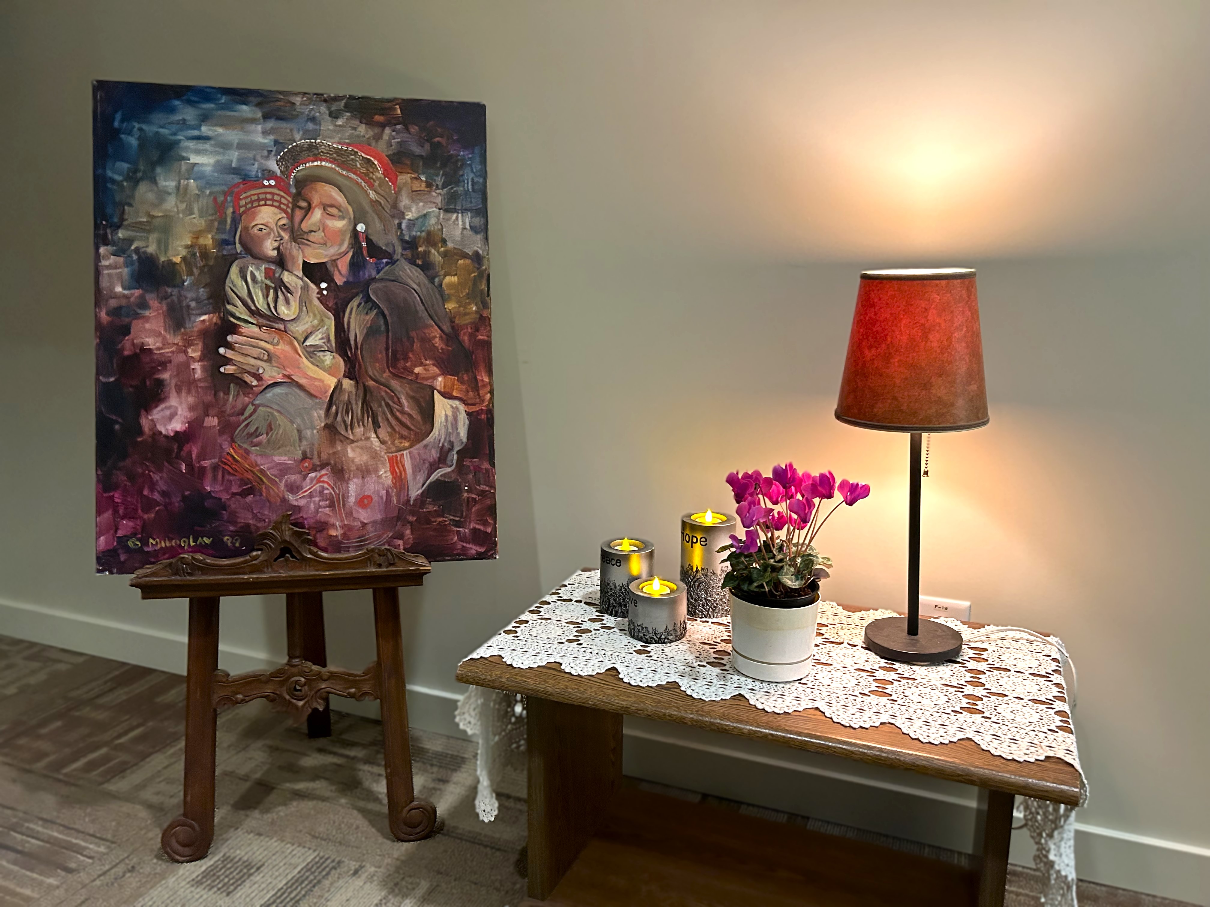 A table set up with a lamp, candles, and a plant. To the left, painted artwork with a mother holding a child.