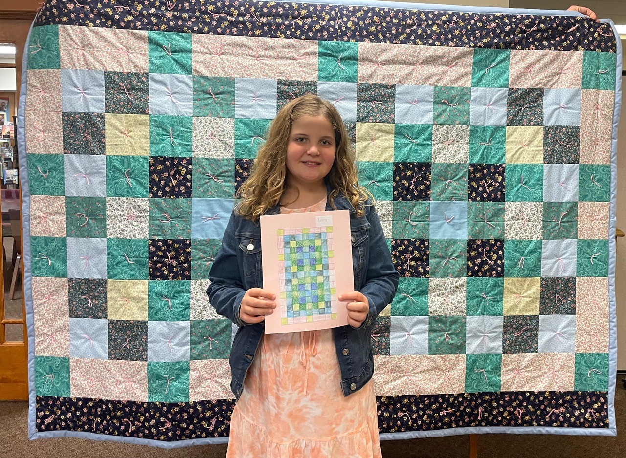 A young girl standing in front of a quilt holding a quilt pattern