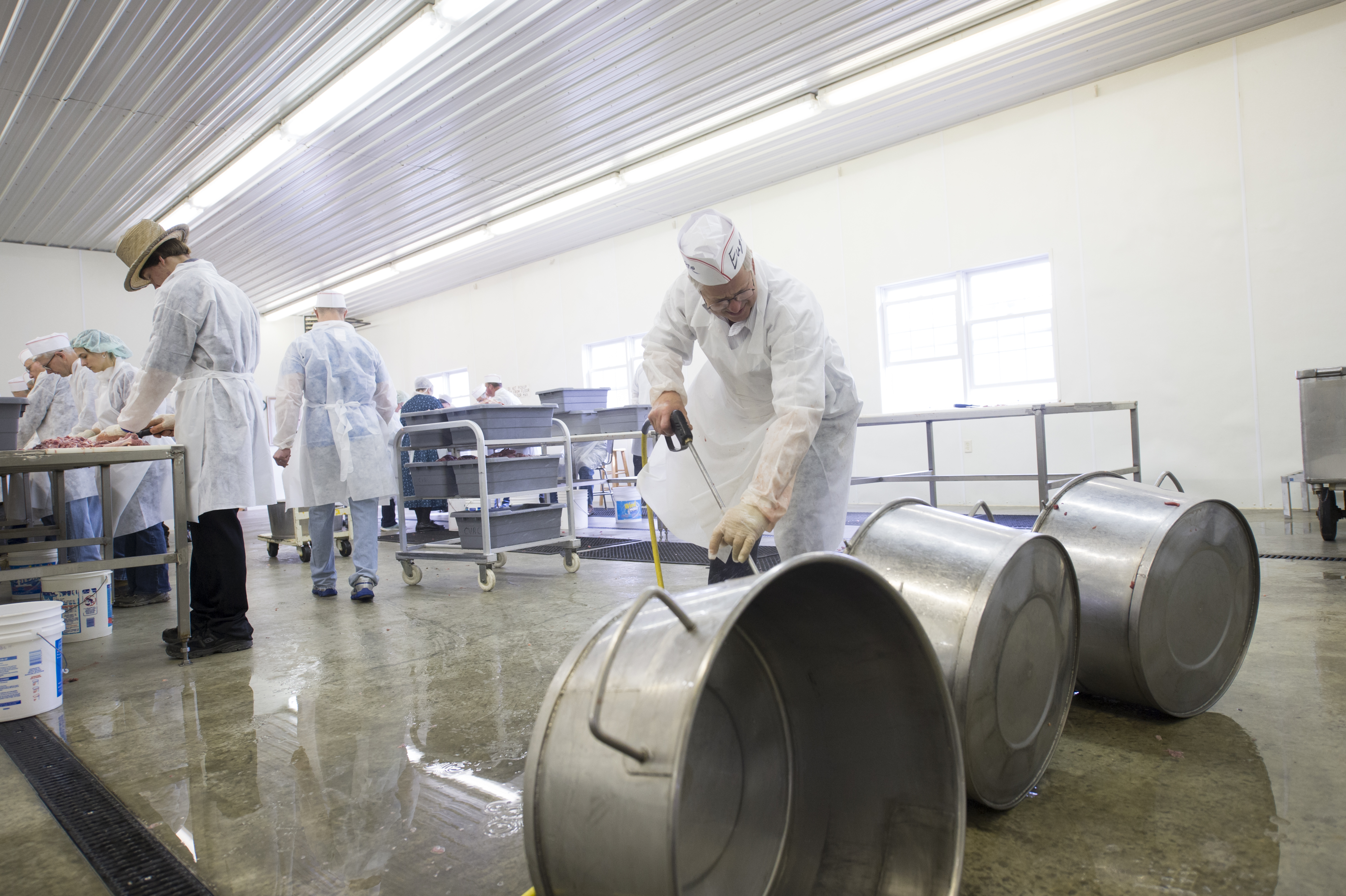 Man sprays off stainless steel tubs while other people in hair nets and aprons work in background
